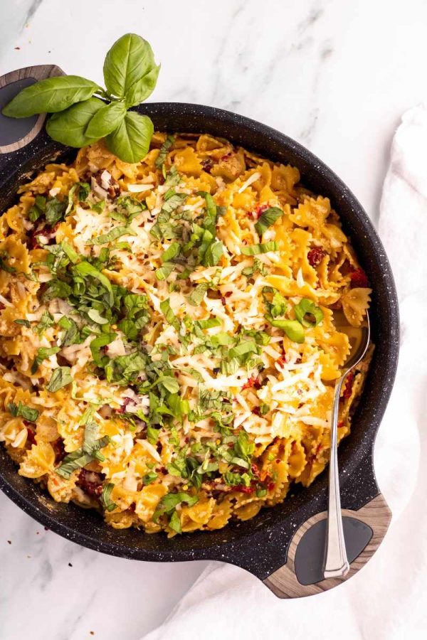 A large serving spoon is tucked into a saucepan of creamy sun-dried tomato pasta, a sprig of fresh basil alongside.