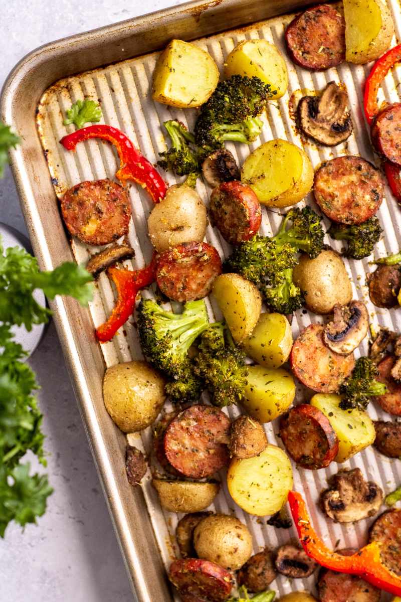 The corner of a metal sheet pan with sausage and veggies rests at an angle alongside fresh parsley.