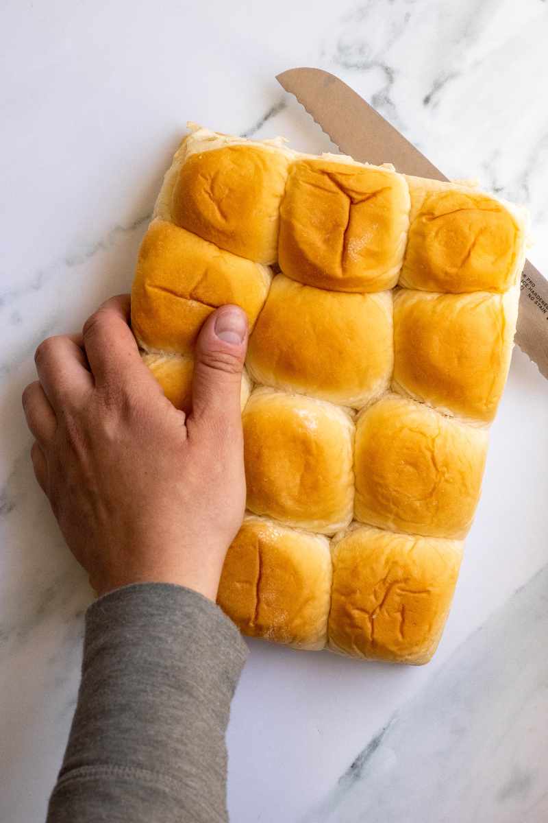 A bread knife slices through twelve slider buns on a white marble counter.