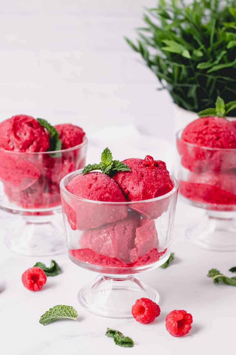 Three footed dessert bowls filled with red frozen dessert on a white counter with fresh mint and berries around.