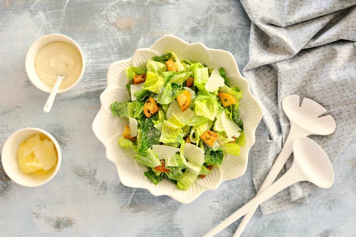 A scalloped bowl with romaine lettuce, croutons, and parmesan shavings with homemade dressing alongside.