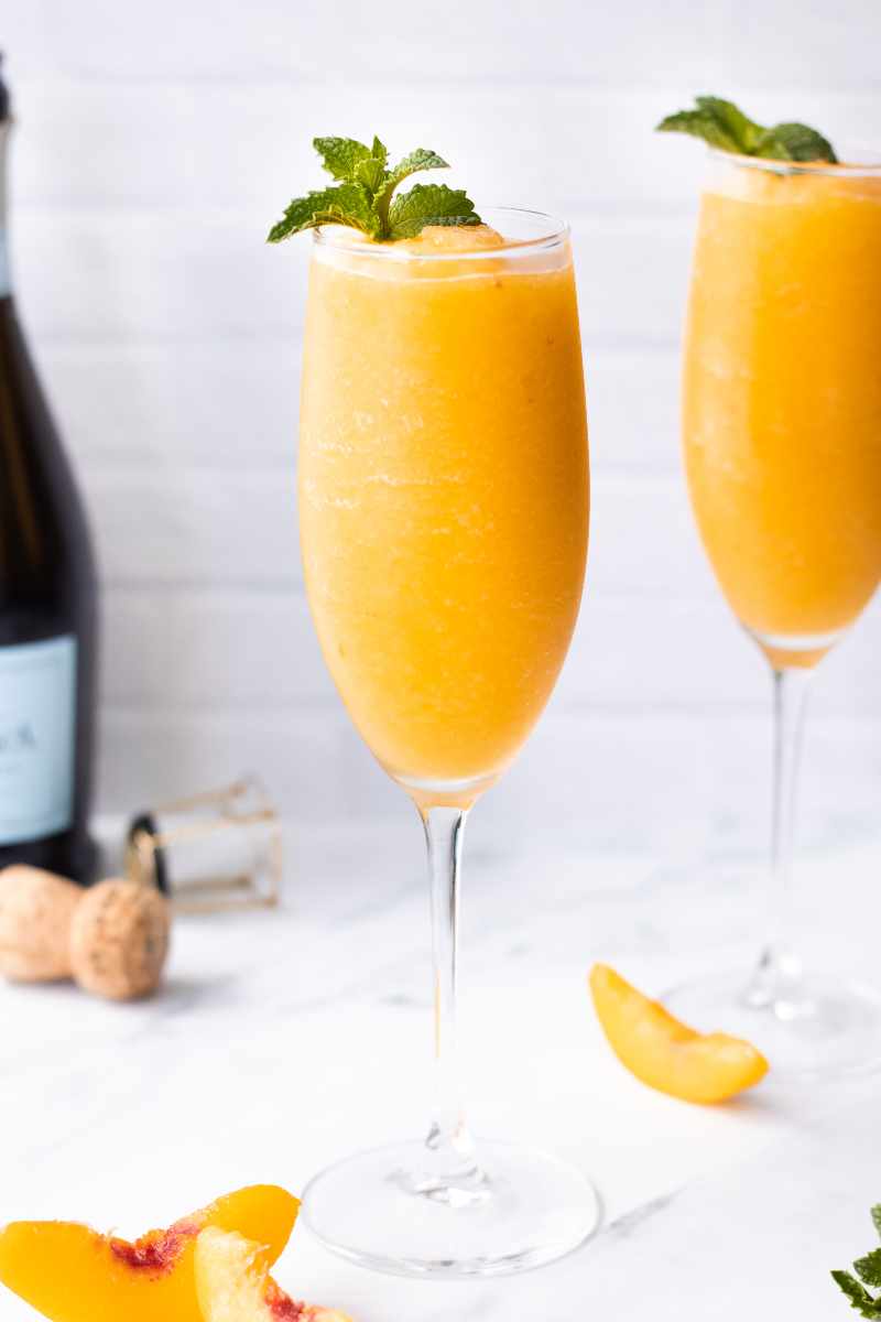 Two peach bellini cocktails in champagne flutes with fresh mint garnish on a white counter.