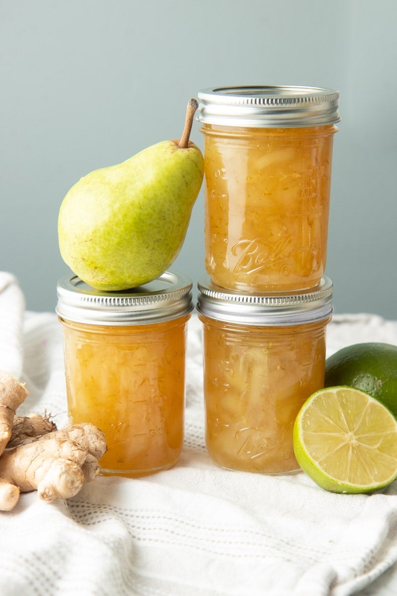 Three half-pint jars of pear preserves and a fresh pear stacked two and two with fresh limes and ginger around.