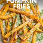 A pile of baked pumpkin fries dusted with chopped herbs and parmesan cheese sits on parchment paper. A text overlay reads, "Baked Parmesan Pumpkin Fries."