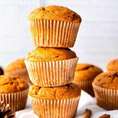 Three tender muffins stacked atop one another with cinnamon sticks and other muffins nearby.