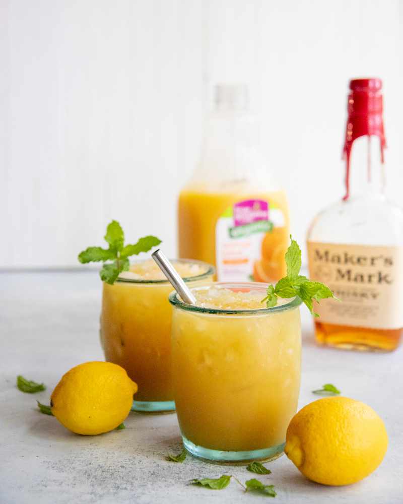 Two bourbon cocktails stand on a light counter surrounded by lemons and mint leaves.