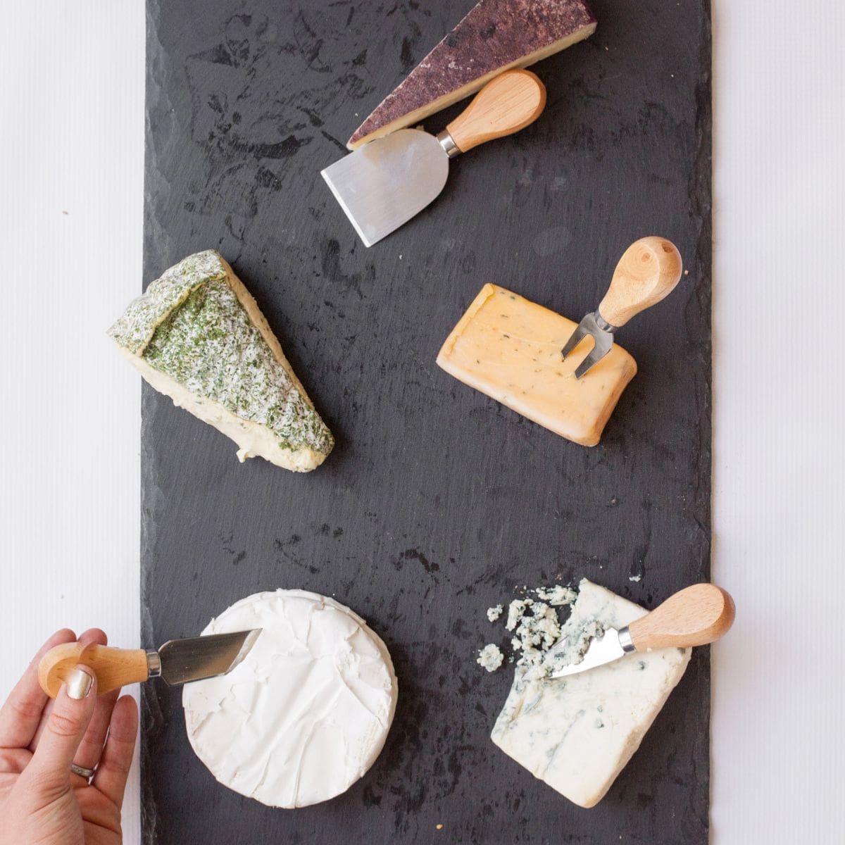 A hand places a cheese knife in wheels of cheese on a cheese tray.