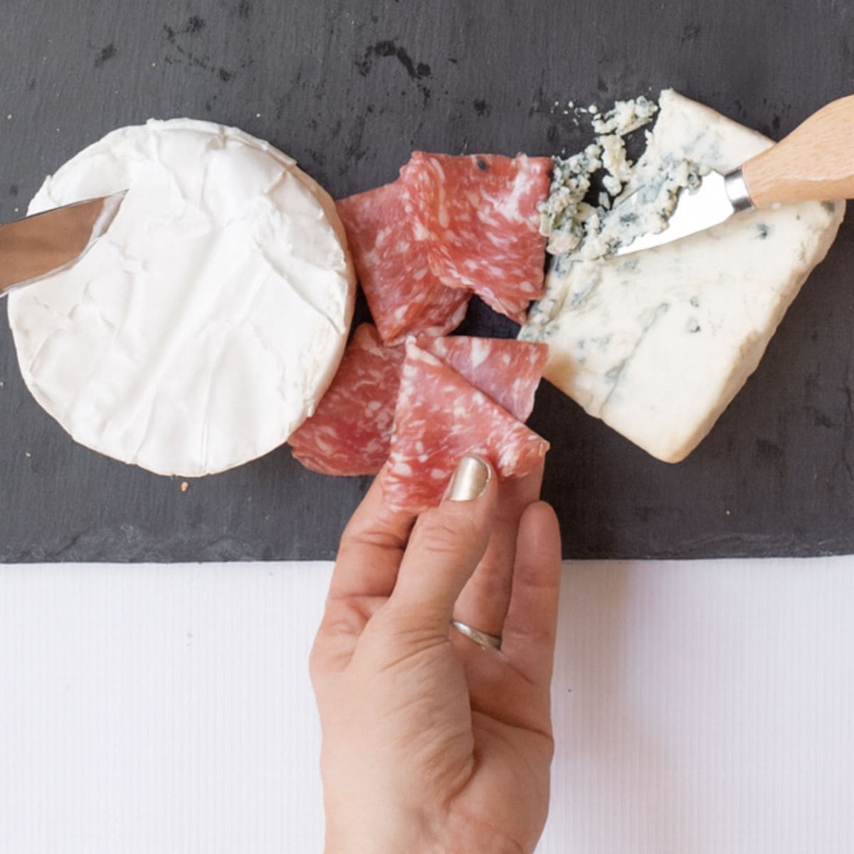 A hand places salami onto a slate cheese board.