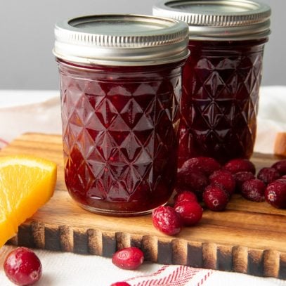 Two quilted half-pint jars of home canned cranberry stand on a wooden board with fresh cranberries and oranges slices around.
