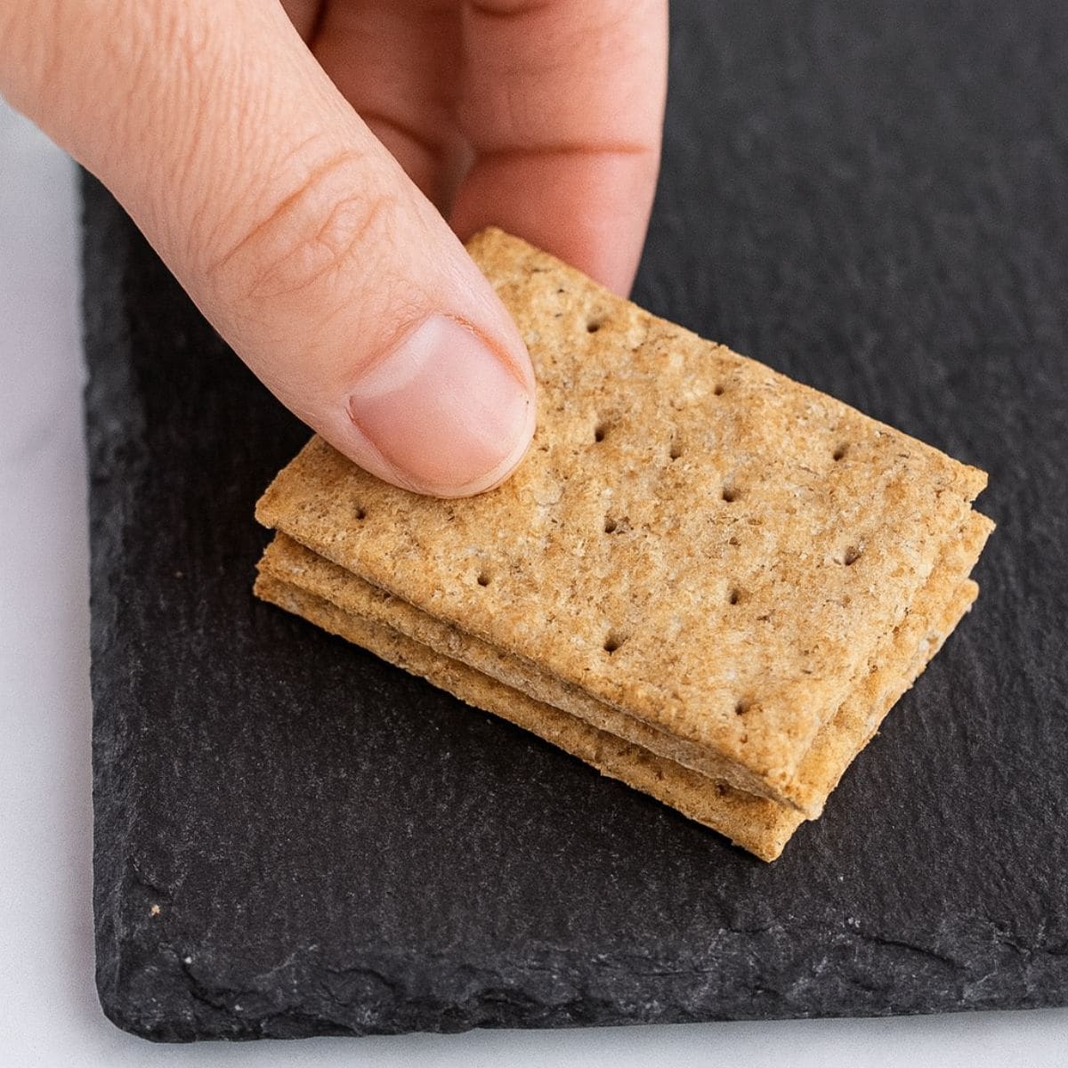 A hand places crackers on the corner of a slate cheese board.
