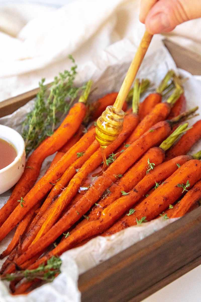 A hand uses a honey dipper to drizzle honey over a pile of oven roasted carrots.