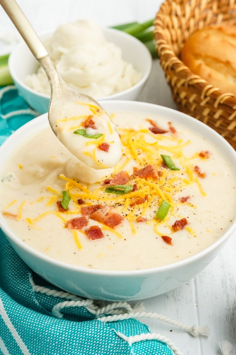 A spoon scoops up a mouthful of soup loaded with toppings such as shredded cheddar cheese, crumbled bacon, and scallions.