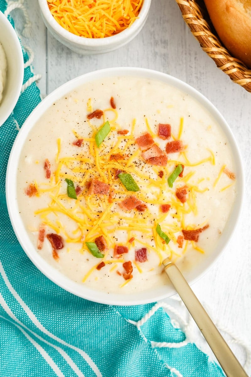 Top view of a bowl of mashed potato soup garnished with shredded cheddar, bacon bits, and green onion.