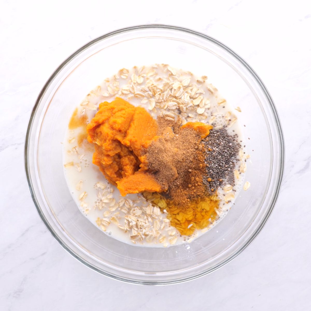 Ingredients for pumpkin overnight oats in a glass bowl on a white marble countertop