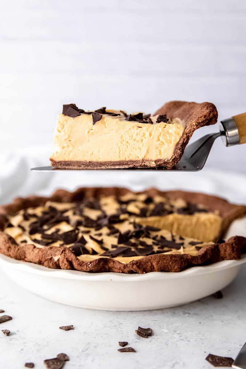 A pie server holds a slice of pie above the garnished pie in a white pie plate.