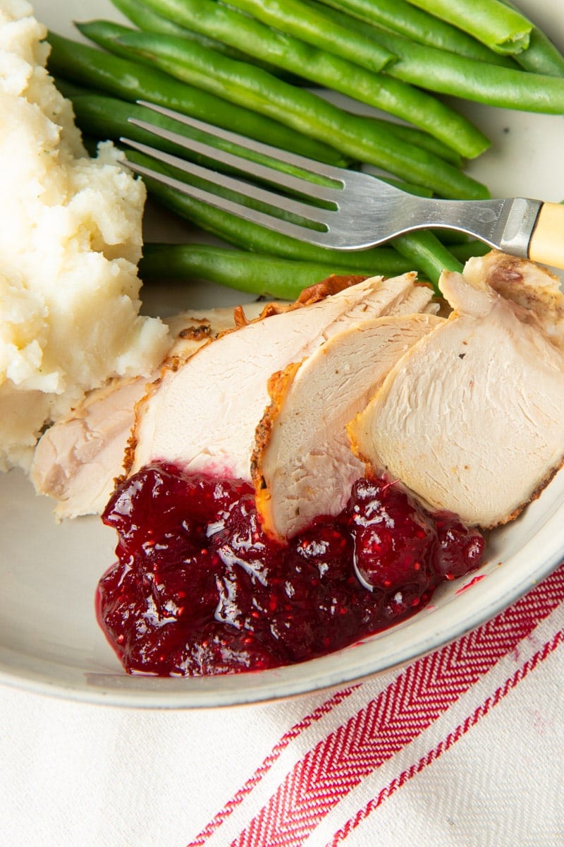 A serving of cranberry sauce on a plate with slices of turkey breast, mashed potatoes, and green beans.