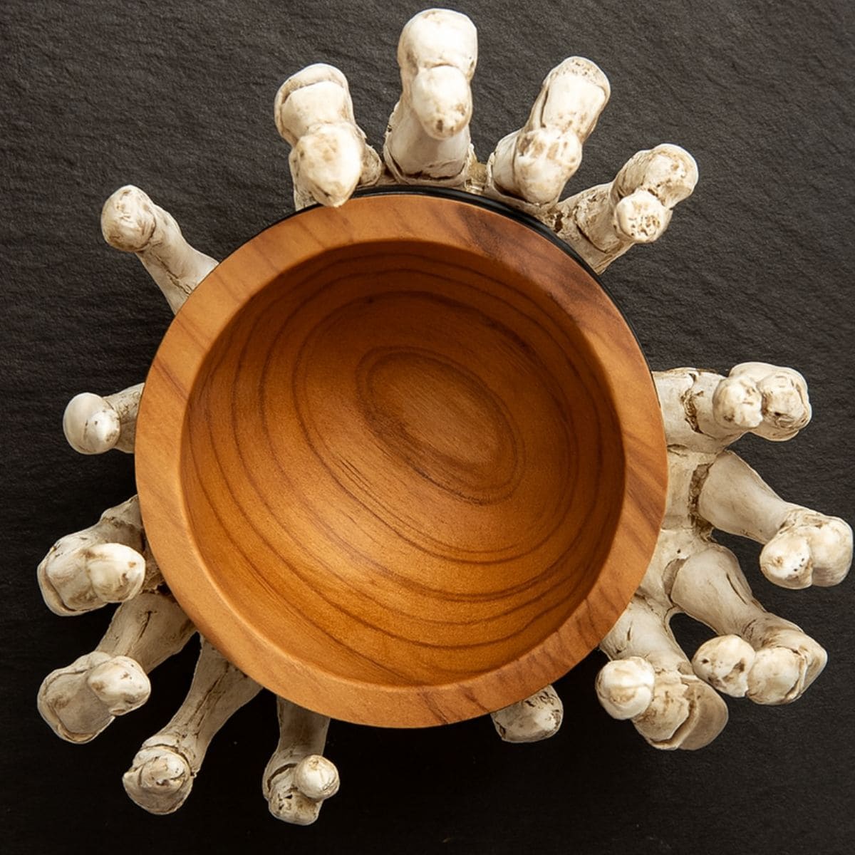 A skeleton hand bowl holder holds a wooden bowl on a black slate board.