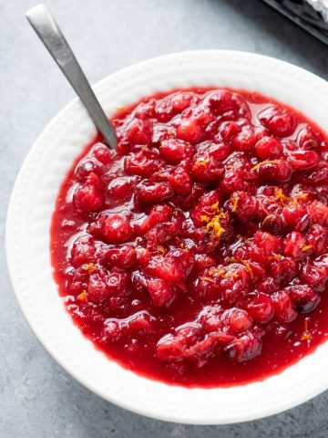 A spoon rests in a white bowl filled with homemade cranberry sauce garnished with citrus zest.