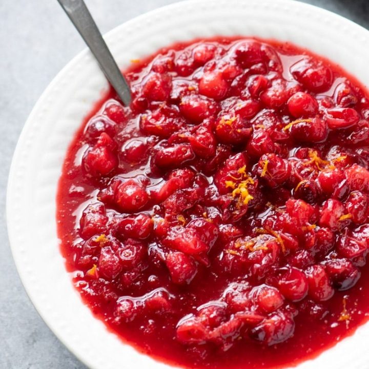 A spoon rests in a white bowl filled with homemade cranberry sauce garnished with citrus zest.