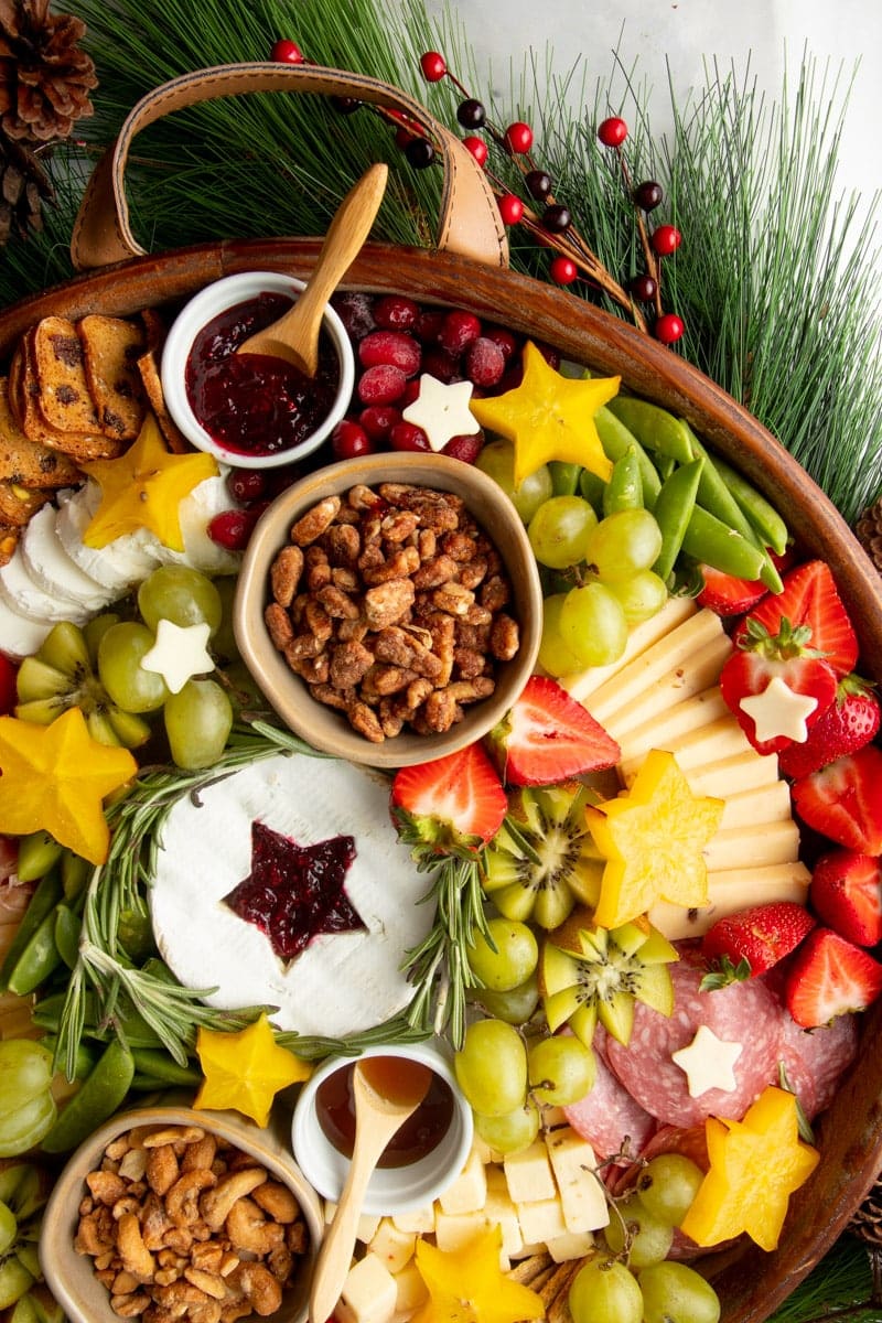 A round Christmas appetizer tray with sliced and cubed cheeses, sliced meats, fresh fruits, bowls of nuts, jams, and honeys, all decorated with star shapes and fresh herbs.