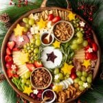 A Christmas charcuterie wreath in a round wooden tray with decorative pine boughs, pinecones, and red berries wreathed around the edges.