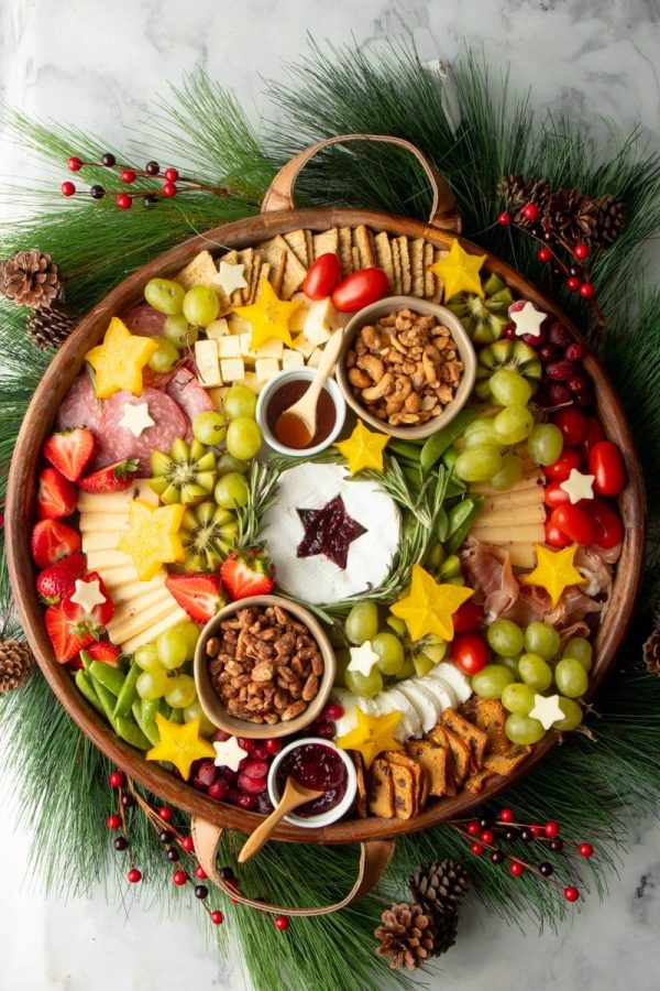 A Christmas charcuterie wreath in a round wooden tray with decorative pine boughs, pinecones, and red berries wreathed around the edges.