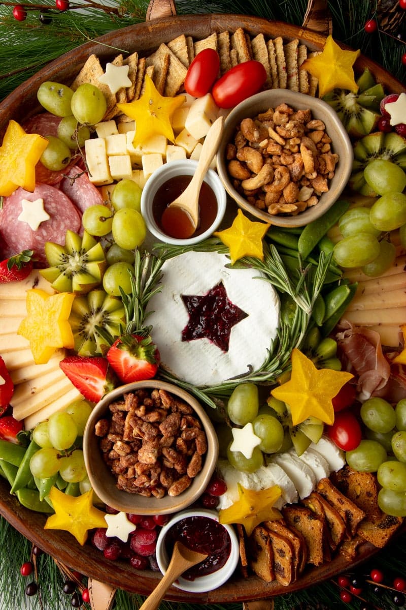 Close view of a Christmas cheese board with a wheel of brie in the center with a star shaped cutout filled with jam, surrounded by additional cheeses, meats, crackers, fruits, and nuts.
