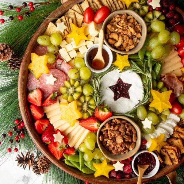 A Christmas snack platter served in a round wooden tray with cheeses, meats, fresh fruits, nuts, jams, and crackers.