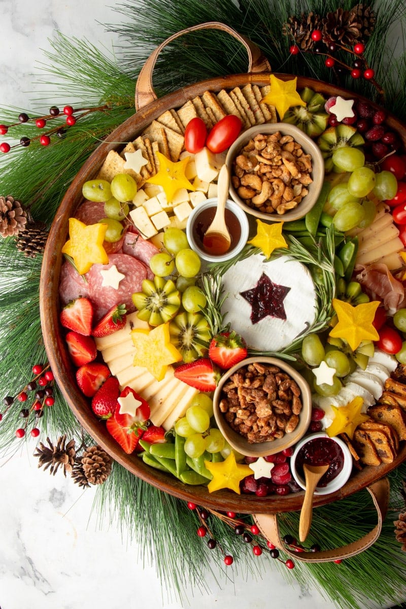 A Christmas snack platter served in a round wooden tray with cheeses, meats, fresh fruits, nuts, jams, and crackers.
