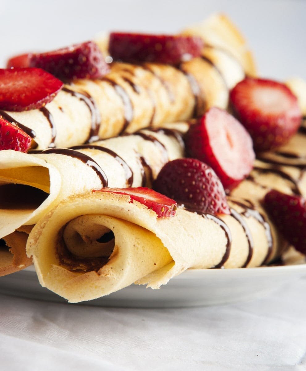 Close view of rolled Dutch pancakes stacked on a plate, drizzled with chocolate sauce, and topped with sliced strawberries.