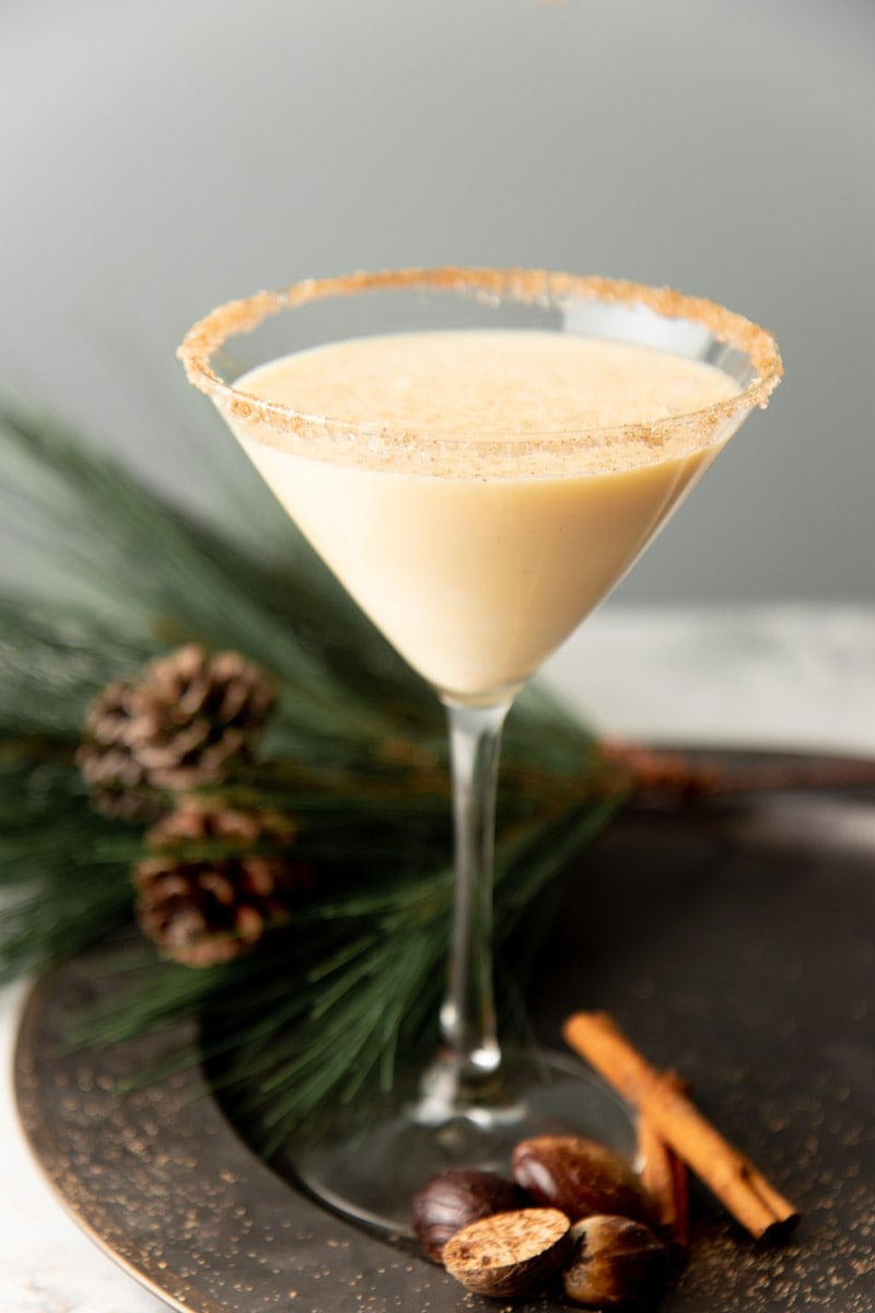 A creamy eggnog martini in a martini glass on a metal serving tray dusted with nutmeg and cinnamon.