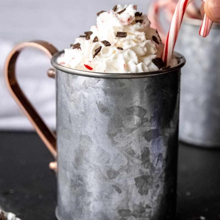 Close view of a whipped cream garnished peppermint mocha in a metal mug as two fingers place a mini candy cane as an additional garnish.