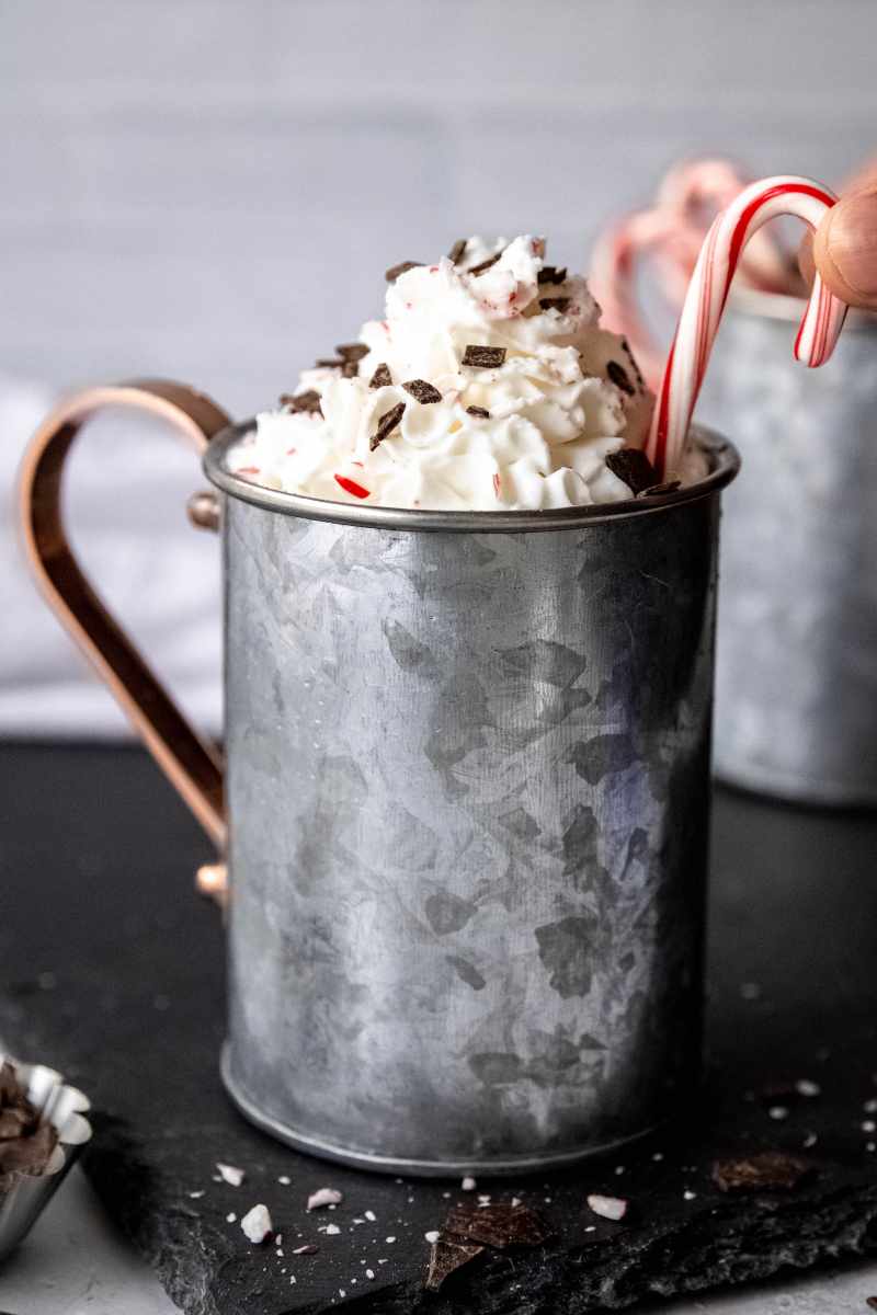 Close view of a whipped cream garnished peppermint mocha in a metal mug as two fingers place a mini candy cane as an additional garnish.