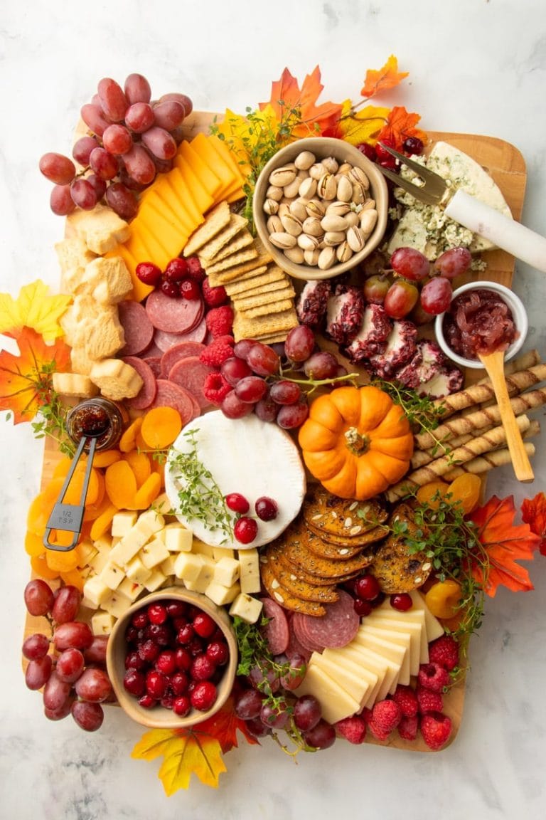 A holiday charcuterie spread decorated for fall with faux leaves, pumpkins, red and orange fruits, and jams.