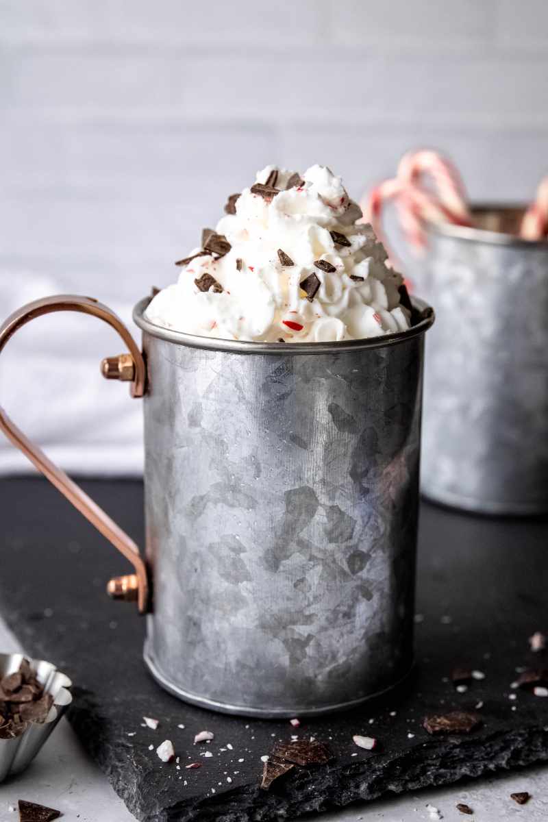 A metal mug with a copper handle filled with homemade peppermint mocha stands on a slate board with a mountain of whipped cream, shaved chocolate, and crushed candy canes as garnish.