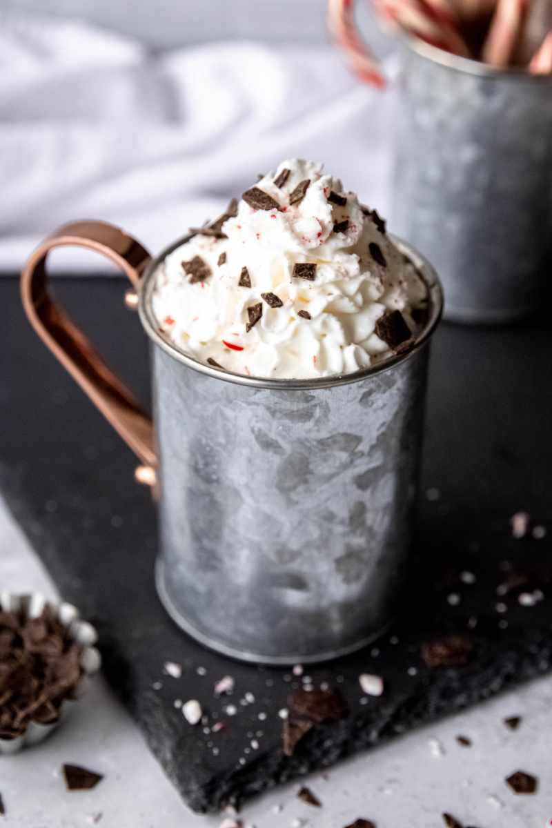 A peak of whipped cream garnished with chocolate shavings and candy cane pieces rises from the top of a metal mug.