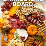 Birdseye view of a wooden cutting board loaded with festive Thanksgiving charcuterie and colorful decorative leaves and mini pumpkins. A text overlay reads, "Festive! Thanksgiving Charcuterie Board."