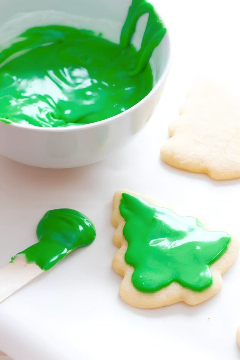 Cookies shaped like evergreen trees sit next to a bowl of green frosting. One cookie has some green icing on it already.