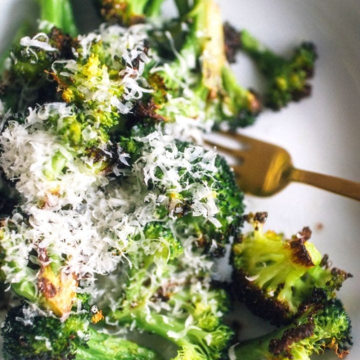Roasted broccoli topped with parmesan cheese on a white serving dish with a gold fork alongside.