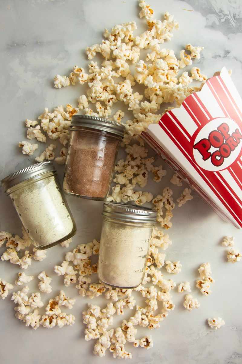 Three jars of homemade seasonings lying on their sides with popcorn spilling from a box all around them.