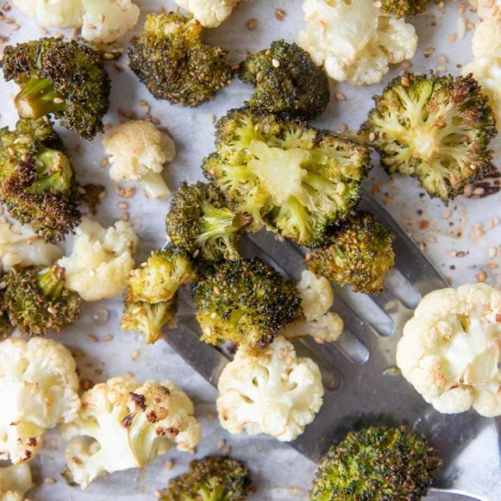 A metal spatula scoops up roasted broccoli and cauliflower from a parchment paper lined sheet pan.
