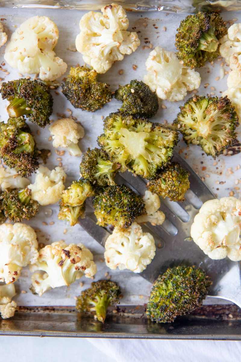 A metal spatula scoops up roasted broccoli and cauliflower from a parchment paper lined sheet pan.
