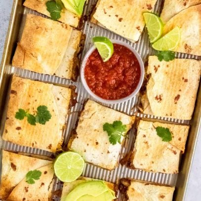 Ground beef sheet pan quesadillas cut into squares on a baking sheet and served with salsa, avocado, and lime.