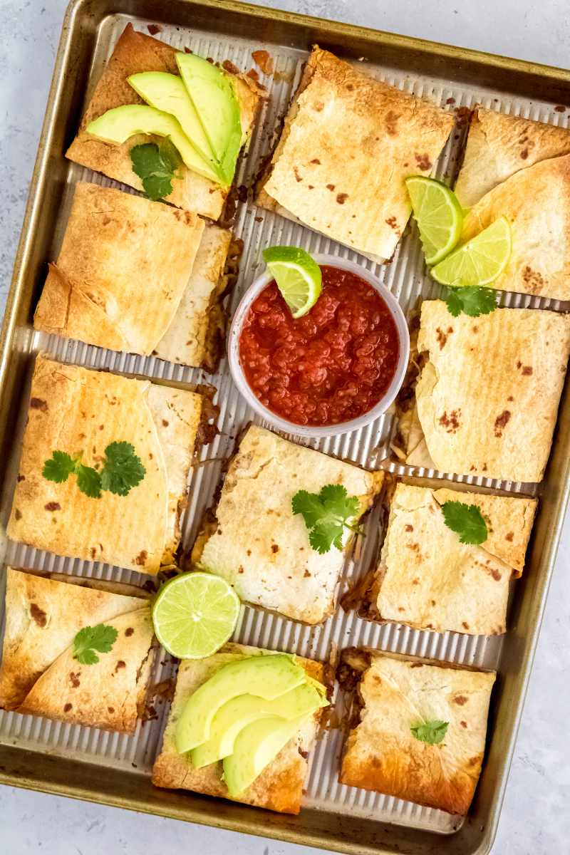 Ground beef sheet pan quesadillas cut into squares on a baking sheet and served with salsa, avocado, and lime.
