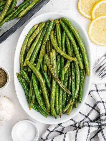 A shallow white dish of oven roasted green beans with additional seasonings around it.