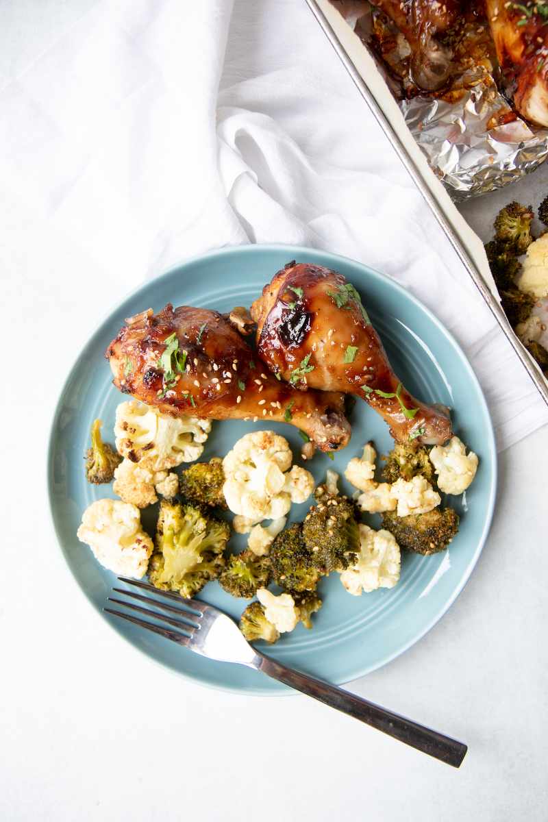 A plate of roasted broccoli and cauliflower with two sticky chicken drumsticks alongside more on a sheet pan.