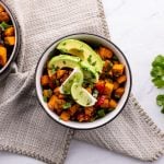 Two black-rimmed bowls filled with sweet potatoes, lime slices, avocado slices, black beans, and rice.