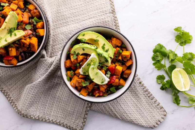 Two black-rimmed bowls filled with sweet potatoes, lime slices, avocado slices, black beans, and rice.