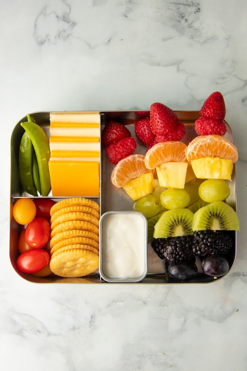 A three-section metal lunch tin with cheese squares and sugar snap peas, grape tomatoes and round crackers, and rainbow fruit kabobs with yogurt dip.