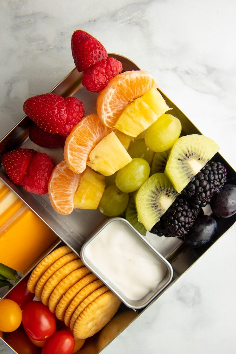 Four rainbow fruit kabobs packed into the large section of a metal bento-style lunch tin with veggies, cheese, and crackers, alongside.